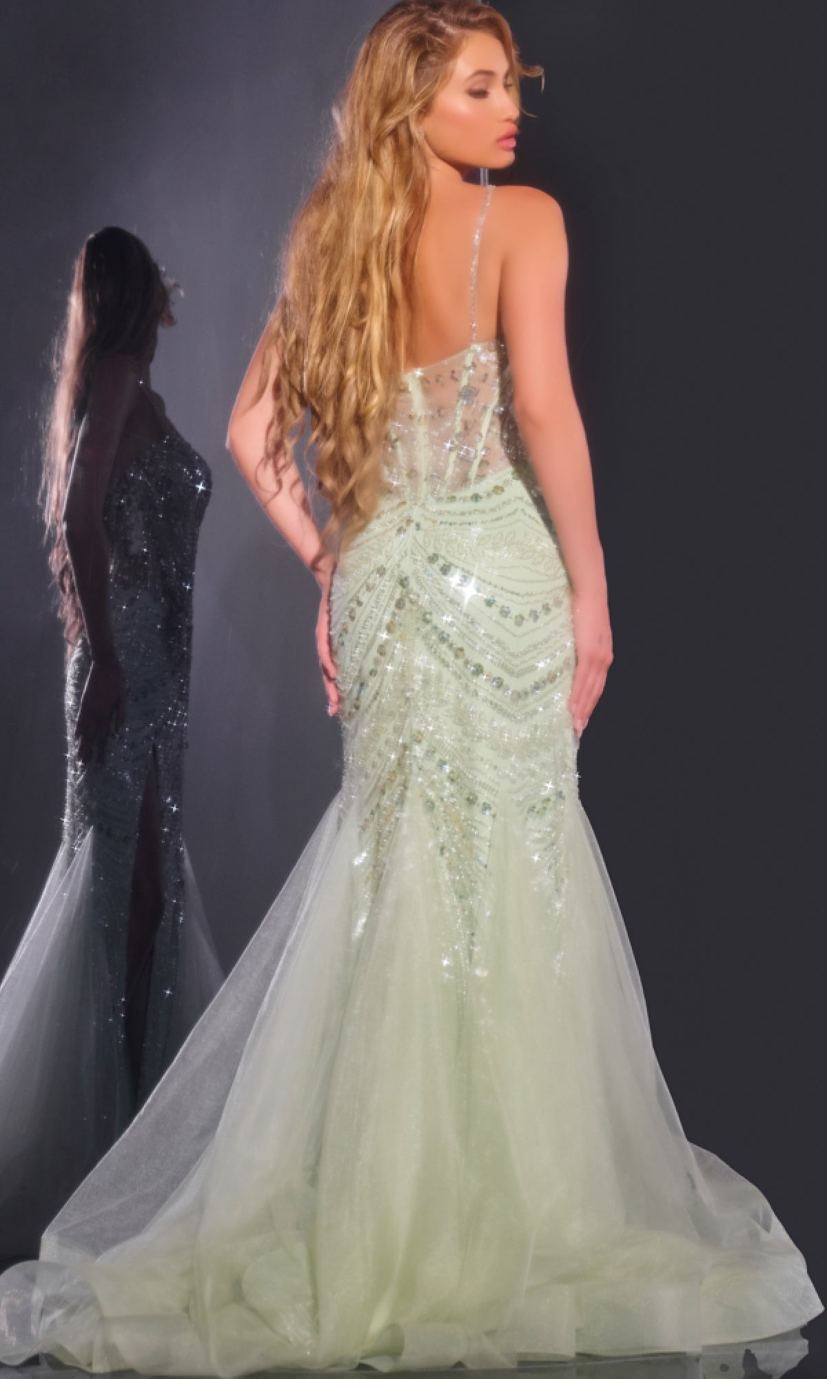 A woman with long wavy hair models the Jovani 43189 long prom dress—a light green, sequined mermaid gown with sparkly embellishments and a tulle train—standing before a mirror and admiring her reflection.